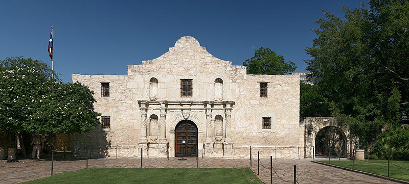 a stone building with a door and a stone fenced yard with Alamo Mission in San Antonio in the background