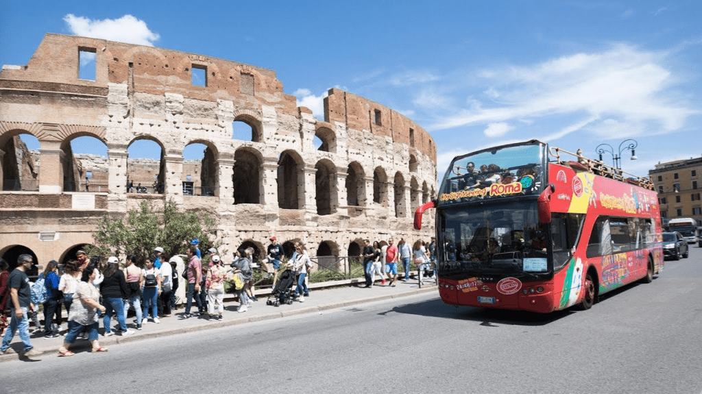 a red double decker bus on a street with people walking around