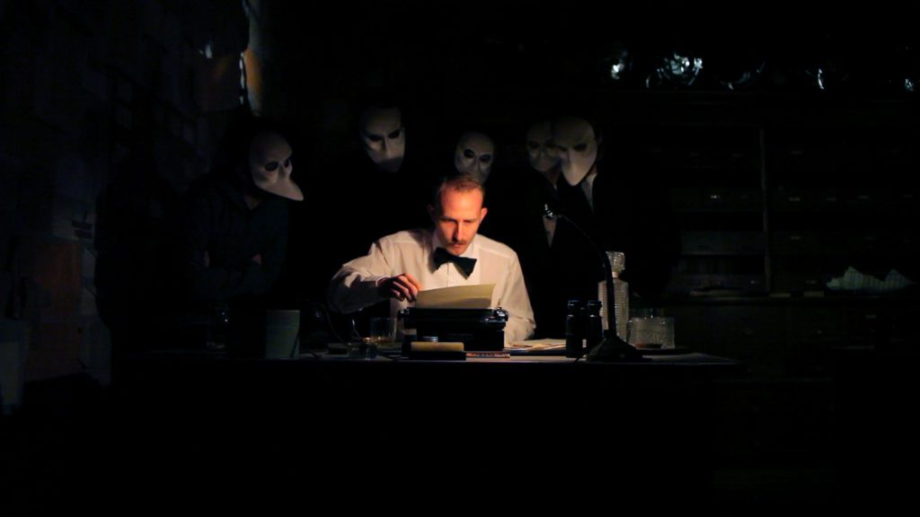 a man in a white shirt and bow tie sitting at a desk with people in masks