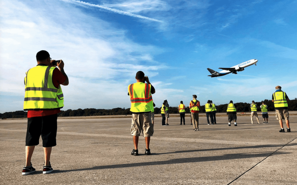 a group of people in yellow vests and a plane flying over a runway