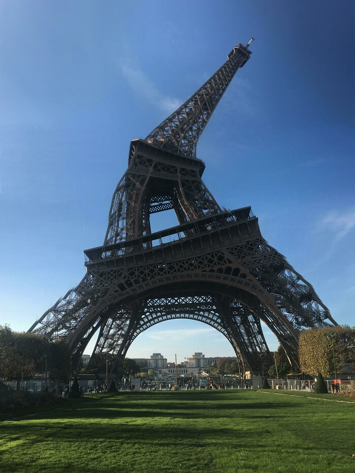 a large metal tower with grass and trees with Eiffel Tower in the background